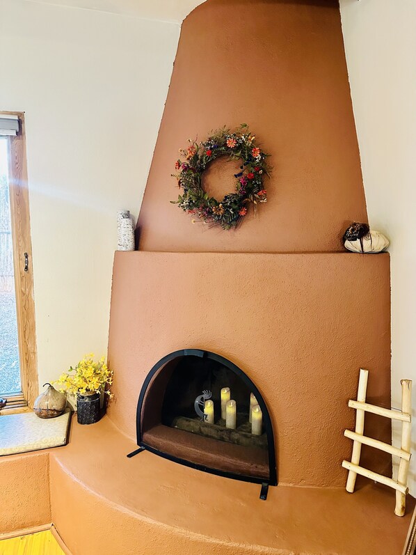 A fireplace with a decorative wreath, a small shelf with decorative items, and a window with a view. - Charming Santa Fe Retreat. Private Patio. 1.4 miles to Plaza. Full kitchen. A/C! (Santa Fe)