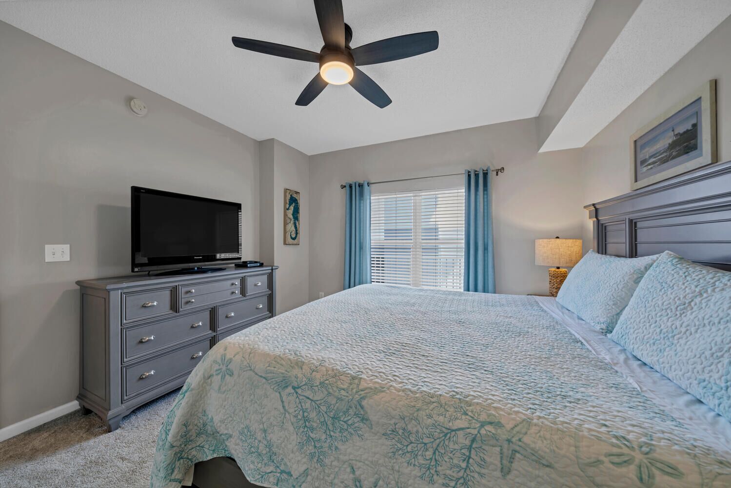 A bedroom with a bed, a dresser, a television, a ceiling fan, and a window with curtains.