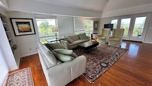 A spacious living room with a beige sofa, a white armchair, and a patterned rug. There are two windows with blinds, a TV, and a small table with a lamp.