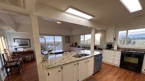 A modern kitchen with white cabinetry, granite countertops, a built-in dishwasher, and a large window offering a view of the outdoors.