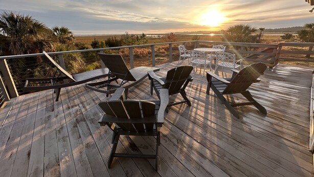 A wooden deck with outdoor furniture, including chairs and a table, overlooking a scenic landscape with a sunset.