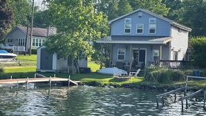 Une maison à deux étages avec une jetée qui s’avance sur une étendue d’eau, entourée de verdure et d’un petit bateau.