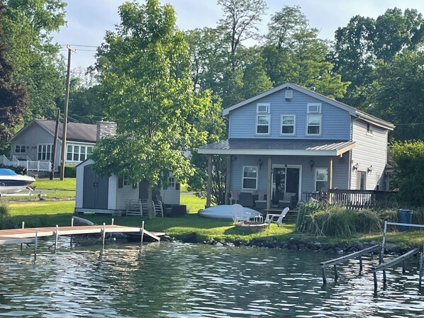 A two-story house with a dock extending into a body of water, surrounded by greenery and a small boat.