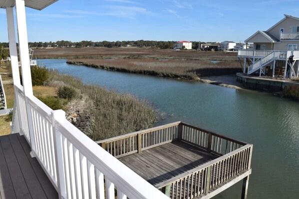 A wooden deck with a railing overlooking a calm river, surrounded by marshland and residential buildings.