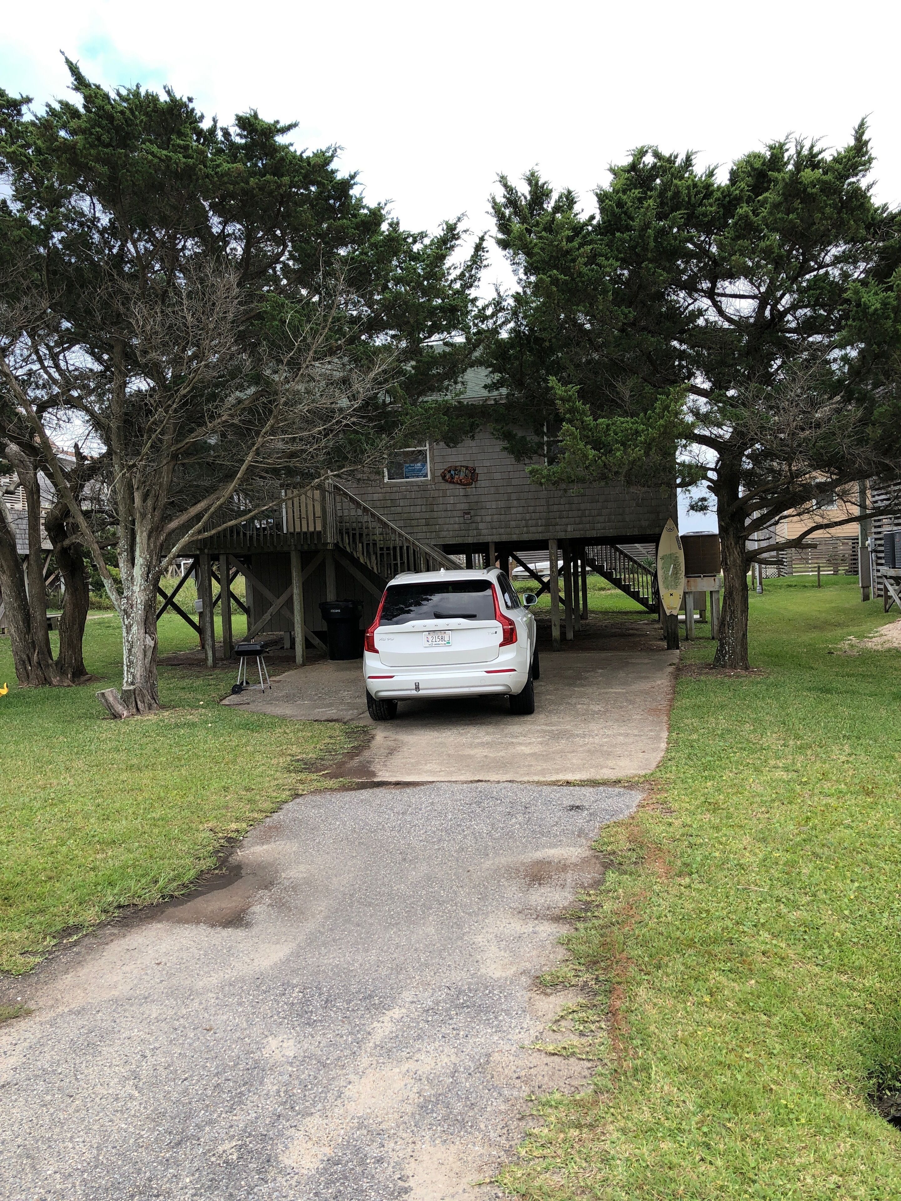 A white car parked under a wooden structure with a covered area, surrounded by trees and a grassy area.