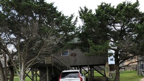 A white car parked under a wooden structure with a covered area, surrounded by trees and a grassy area. - Spectacular Sound & Ocean Views, OBX 3BR2BA House, sleeps 8, dog friendly (Frisco)