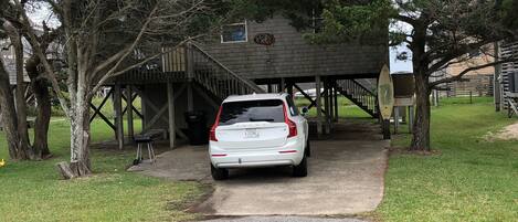 A white car parked under a wooden structure with a covered area, surrounded by trees and a grassy area.