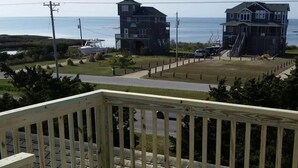 A wooden deck with a railing, overlooking a beach with two buildings and a boat in the background. - Spectacular Sound & Ocean Views, OBX 3BR2BA House, sleeps 8, dog friendly (Frisco)