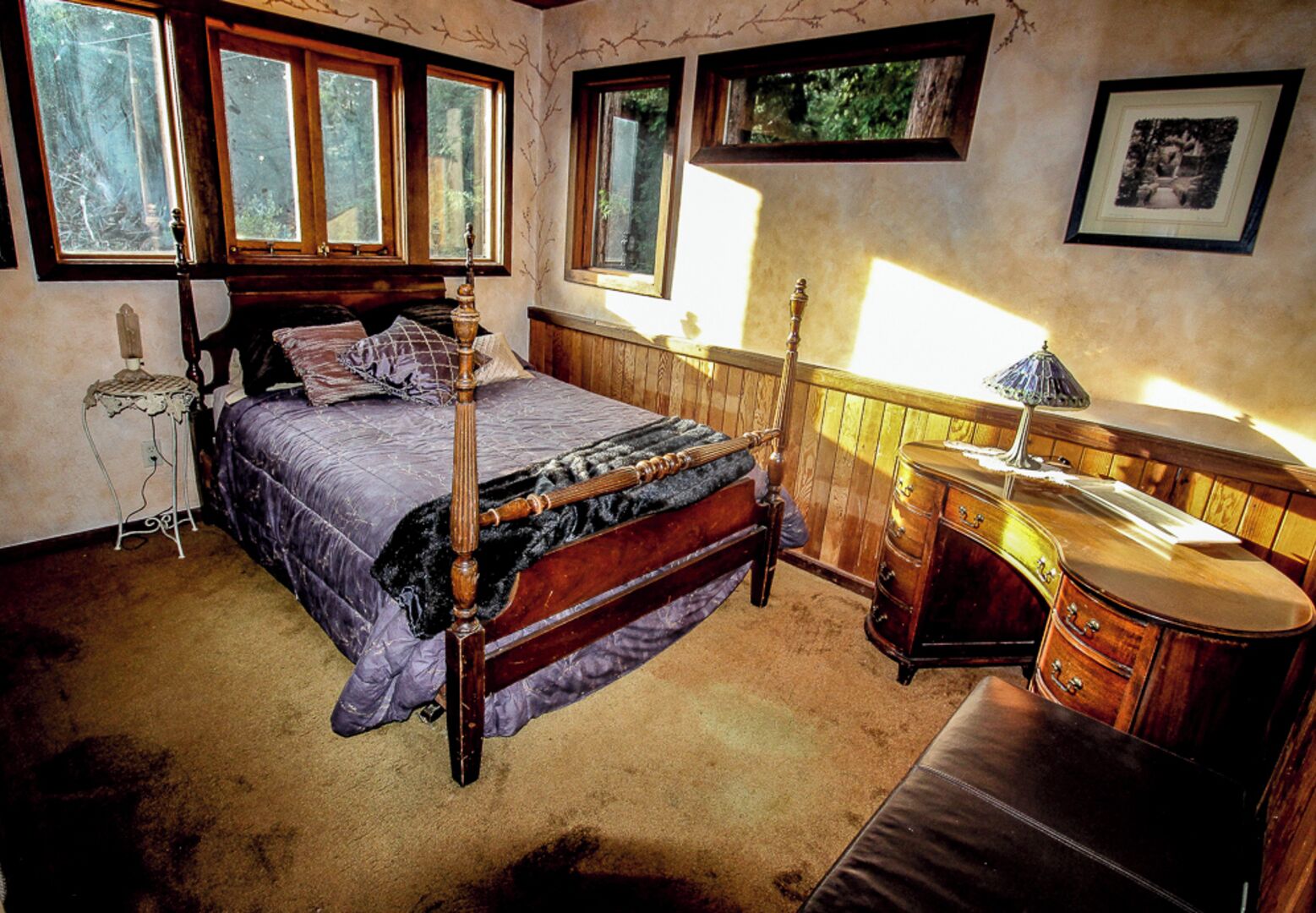 A bedroom with a wooden four-poster bed, a dresser, and a nightstand. There are large windows allowing natural light.