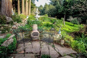 Relax in a serene garden setting surrounded by lush greenery and a stone pathway leading to a seating area with a stone oven.