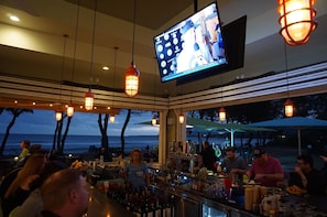 Una zona de bar con una pantalla de televisión, luces colgantes y vistas al océano.