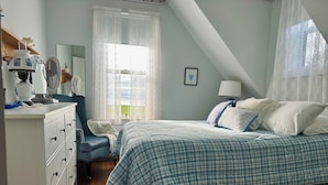 Un dormitorio con cama, cómoda, silla y ventana con cortinas de encaje, y vistas al exterior.