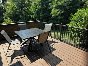 An outdoor deck with a table and chairs, surrounded by a metal railing and dense foliage.