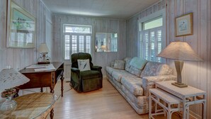 Una habitación con piso de madera, un sillón verde, un sofá estampado, una mesita y una lámpara.