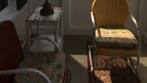 Une pièce avec une fenêtre, une lampe sur une petite table, une chaise à motifs floraux et une chaise avec un cadre en métal.