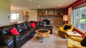 A living room with a black leather sofa, a round coffee table, and a yellow armchair. There is a brick fireplace and a window with curtains.