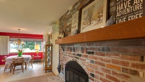 A cozy living room with a brick fireplace, a dining area with a table and chairs, and a window with curtains.