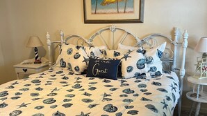 A bed with a white and blue seashell and starfish pattern, a framed painting above it, and a nightstand with a lamp and a book. - Perfect Key West Style - Golf -Tennis - Pet Friendly (Bethany Beach)