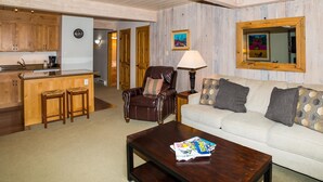 A living room with a sofa, armchair, coffee table, and a small kitchen area with a bar stool. - Lift One - Rustic second floor one bedroom with balcony (Aspen)