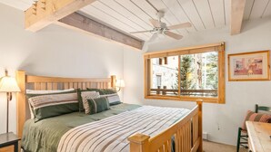 A bedroom with a wooden bed, a window with a view, a ceiling fan, and a nightstand with a lamp. - Lift One - Rustic second floor one bedroom with balcony (Aspen)