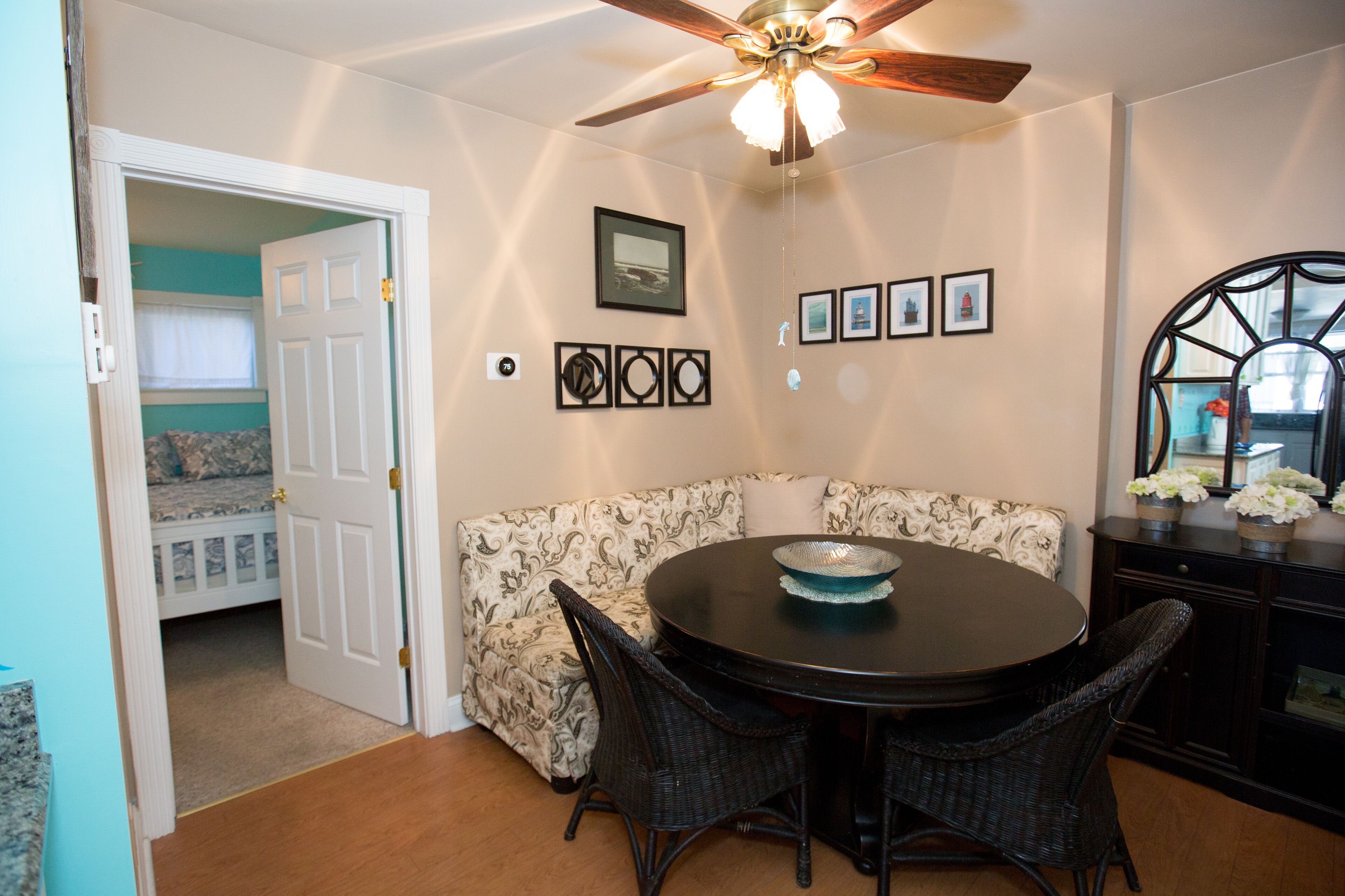 A dining area with a round table, wicker chairs, a ceiling fan, and a wall with framed pictures.