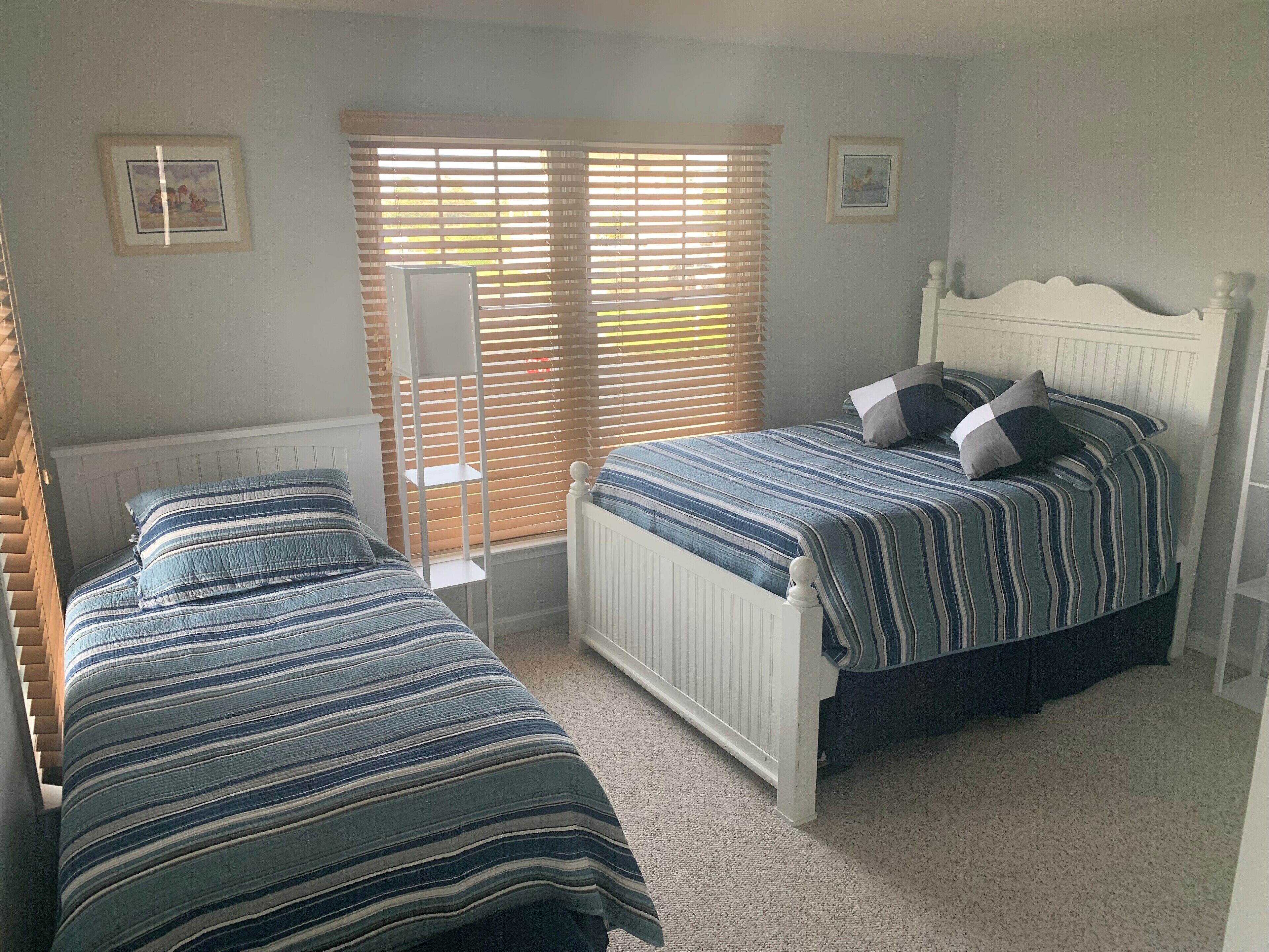Une chambre avec deux lits simples, un radiateur, une fenêtre avec des stores et deux tableaux encadrés accrochés au mur.