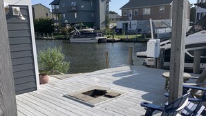 Een houten terras met Adirondack-stoelen, een vuurkorf en uitzicht op een gracht met boten en huizen op de achtergrond.