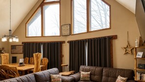 A cozy living room with a high ceiling, a dining area with wooden chairs, and a bookshelf.