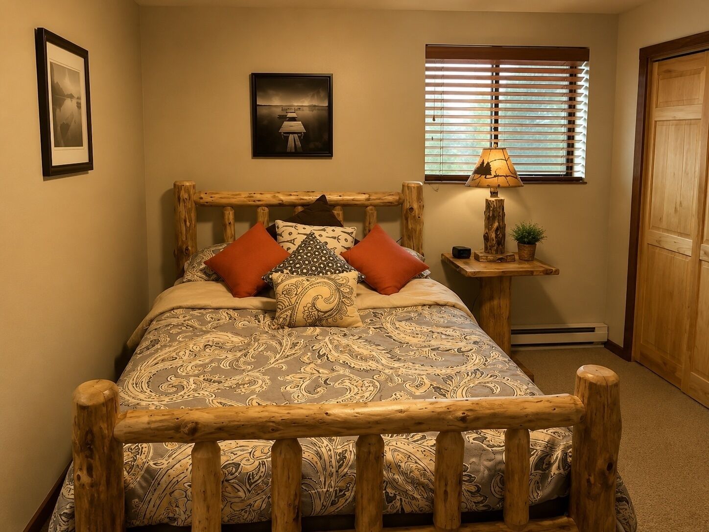Un lit avec une tête de lit en bois, une table de chevet avec une lampe, un tableau encadré au mur et une fenêtre avec des stores.