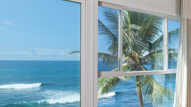 A room with a large window overlooking a beach with waves and palm trees. - Oceanfront Penthouse, As Close As You Can Stay To Ocean, Stunning Views, A/C! (Kailua-Kona)