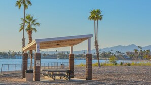Un pavillon situé au bord de la plage, avec un banc et une poubelle, d’où l’on peut admirer un plan d’eau bordé de palmiers et dominé par des montagnes en arrière-plan.