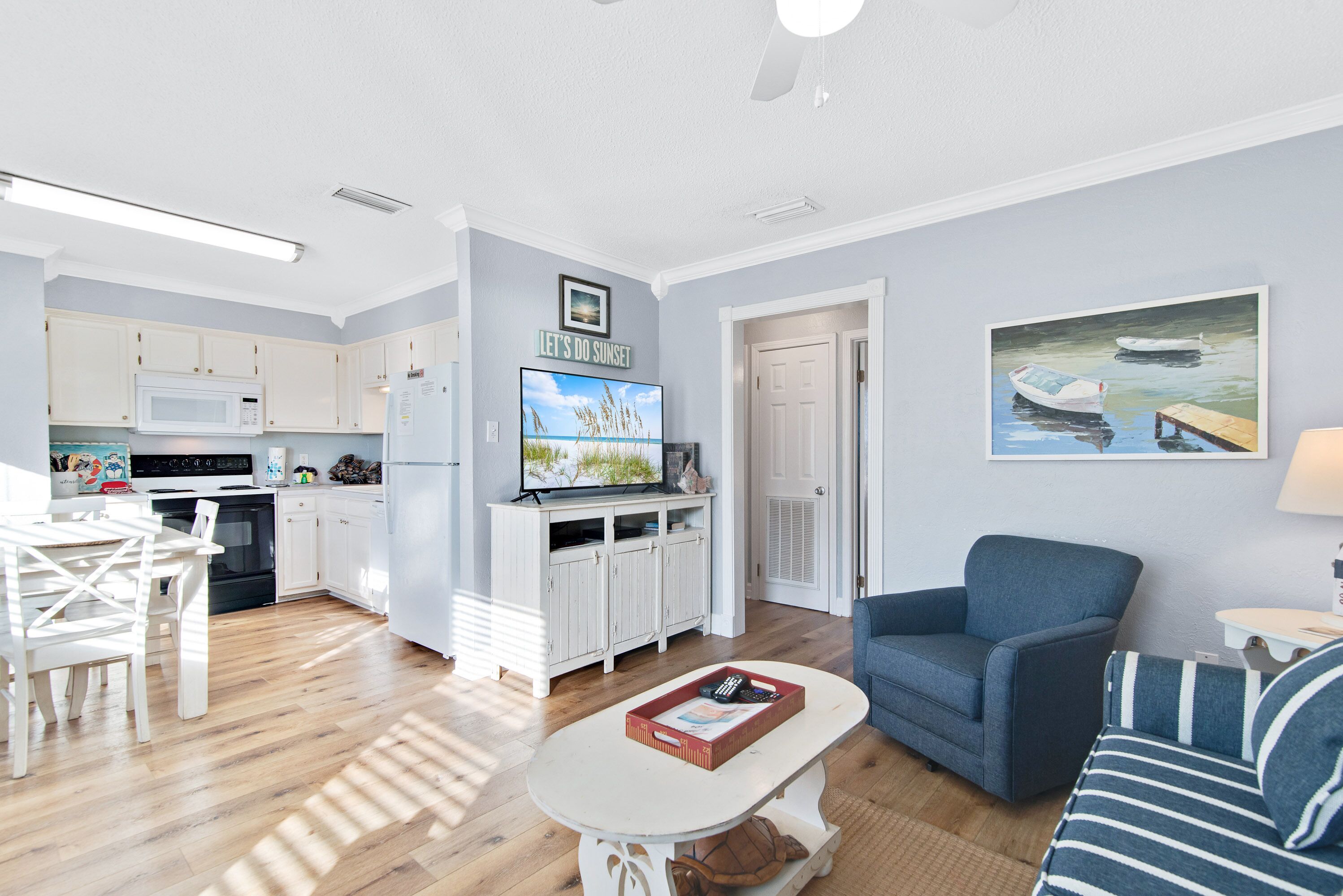 A modern kitchen with white cabinets, a microwave, and a dining area with a white table and chairs. There is a living room with a blue sofa and a painting of boats on the wall.