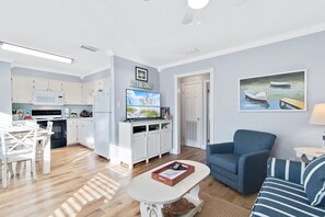A modern kitchen with white cabinets, a microwave, and a dining area with a white table and chairs. There is a living room with a blue sofa and a painting of boats on the wall.