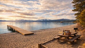 A beach with a wooden dock and picnic table with wine glasses and a basket.