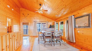 A wooden cabin interior with a dining area featuring a table and chairs, a ceiling fan, and a large window with curtains. - Heavenly View Cabin (Sevierville)