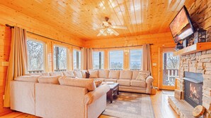 A cozy log cabin interior with a large L-shaped sectional sofa, a fireplace, and a television mounted on the wall. - Heavenly View Cabin (Sevierville)