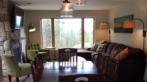 A dining area with a wooden table and chairs, a stone fireplace, a television, and a large window with a view of trees. - Ski In/Out, Walk To Trails & Town, 4 Bdr Townhome. (Breckenridge)