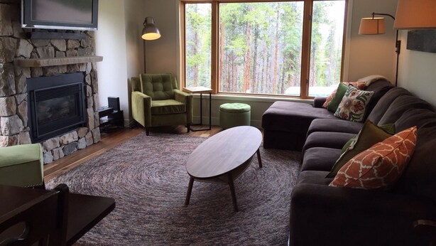 A living room with a stone fireplace, a green armchair, a sofa with patterned cushions, a wooden coffee table, and a large window with a forest view. - Ski In/Out, Walk To Trails & Town, 4 Bdr Townhome. (Breckenridge)