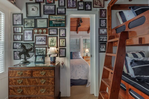 Een slaapkamer met een stapelbed, een dressoir met lades, een ventilator en een wand versierd met ingelijste foto's.