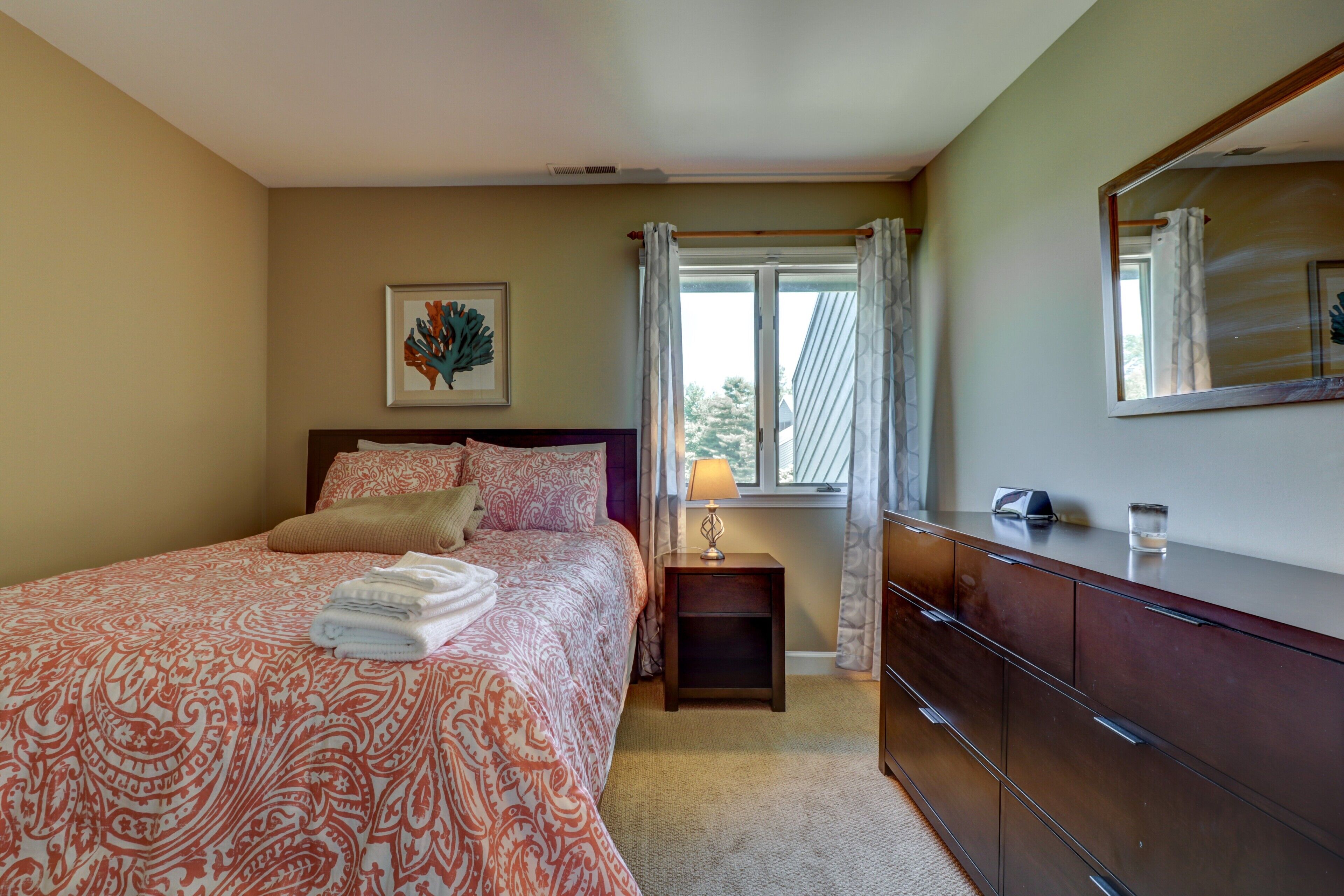 Une chambre à coucher comprenant un lit, une table de chevet, une commode, une fenêtre avec des rideaux et un tableau encadré accroché au mur.