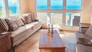 A living room with a beige sofa, a wooden coffee table, and a view of the ocean through large windows. - Gorgeous, Spa-like 3BR with Island Elegance & Stunning Ocean Views (St Thomas)