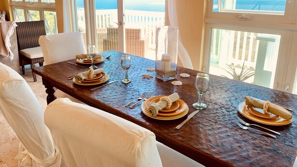 A dining table set for four with white chairs, a view of the sea through large windows, and a balcony with wicker furniture. - Gorgeous, Spa-like 3BR with Island Elegance & Stunning Ocean Views (St Thomas)