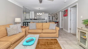 A living room with a sofa set, a coffee table, and a remote control. There is a ceiling fan, a lamp, and a door leading to another room. - Come on down! Great weather, comfortable furniture and beds! 
Best view! (Surfside Beach)