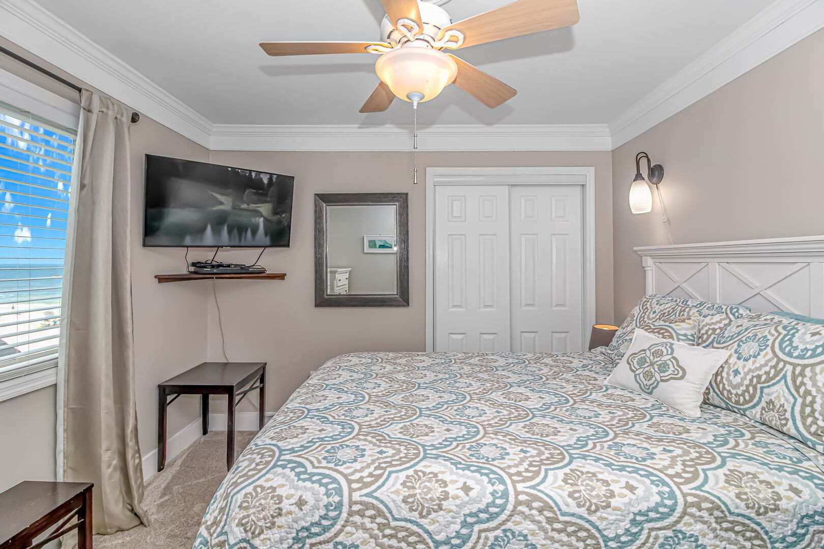 Une chambre à coucher avec un ventilateur de plafond, une télévision, un lit avec une literie à motifs, une table de chevet et une commode.