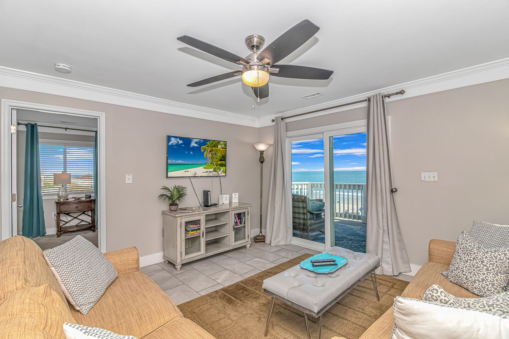 A living room with a ceiling fan, a large window with a view of the beach, a TV mounted on the wall, and a small cabinet.