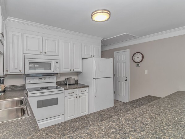 A kitchen with white cabinets, a microwave, a stove, a toaster, a refrigerator, granite countertops, and a clock on the wall.