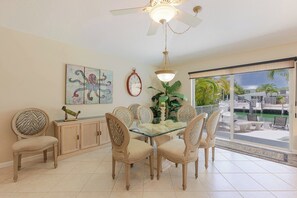 A dining area with a glass table, beige chairs, a ceiling fan, a wall clock, and a large window with a view of a pool and palm trees.