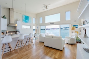 A modern kitchen with white cabinets, a dining area with white chairs, a white sofa, and a view of the water.