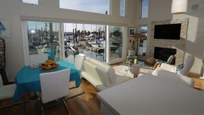 Un vaste espace de vie avec une grande porte vitrée donnant sur une vue du port de plaisance, un téléviseur à écran plat fixé sur un mur en pierre et un coin repas avec une nappe bleue.