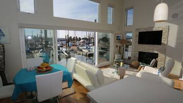 Un vaste espace de vie avec une grande porte vitrée donnant sur une vue du port de plaisance, un téléviseur à écran plat fixé sur un mur en pierre et un coin repas avec une nappe bleue.